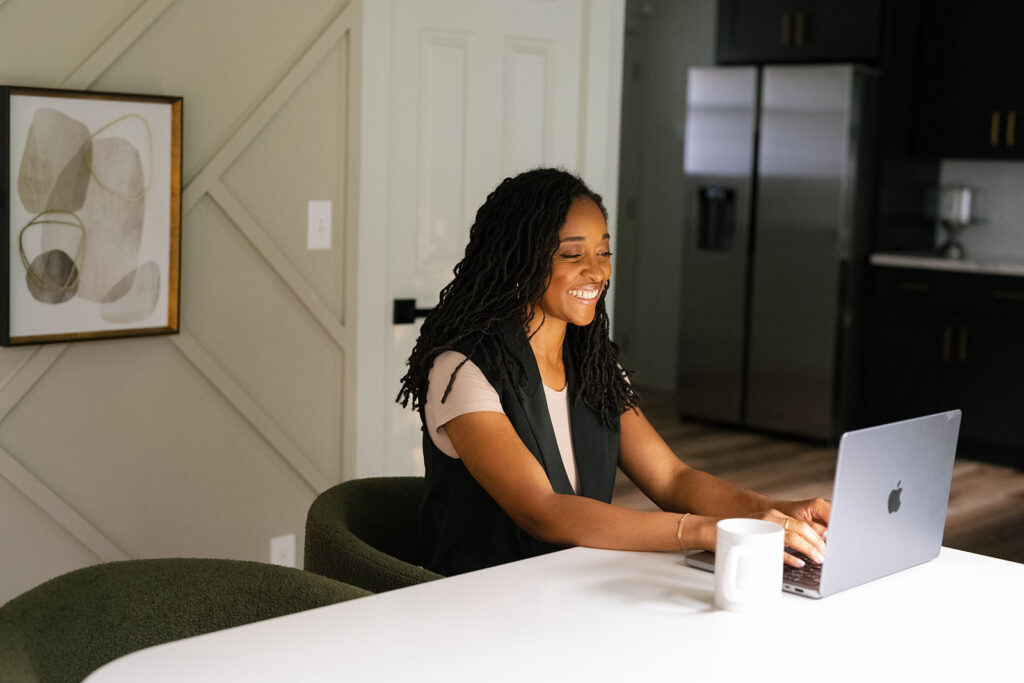 Imani fitch, Founder of Confident Care Birth sits at a modern table, using a laptop and holding a white mug, in a stylish kitchen setting.