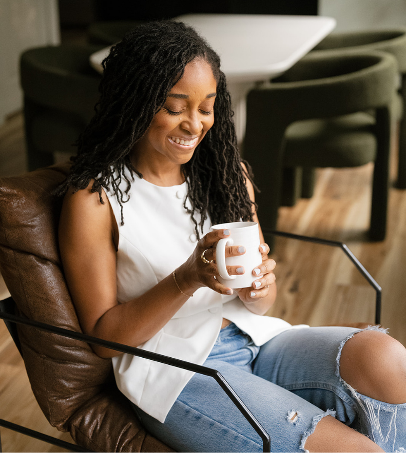Imani fitch, founder of Confident Care Birth with long hair in a white sleeveless top and ripped jeans, smiling while holding a white mug, seated on a brown chair in a cozy room.